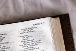 An open book displaying text from Psalms 34:19, with a focus on the verse and surrounding text. The book's cover is partially visible, and there is soft lighting, suggesting a calm or introspective setting.