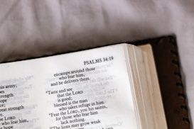 An open book displaying text from Psalms 34:19, with a focus on the verse and surrounding text. The book's cover is partially visible, and there is soft lighting, suggesting a calm or introspective setting.