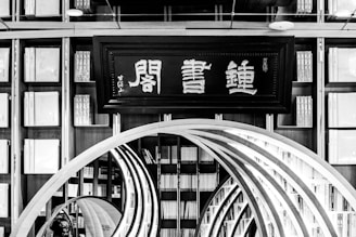 An interior view of a library featuring a large traditional wooden bookshelf filled with numerous books. At the center is a decorative wooden archway in the form of concentric arches leading deeper into the space. Above the archway, there is a framed sign displaying Asian characters.