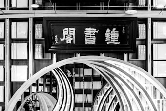 An interior view of a library featuring a large traditional wooden bookshelf filled with numerous books. At the center is a decorative wooden archway in the form of concentric arches leading deeper into the space. Above the archway, there is a framed sign displaying Asian characters.