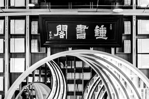 An interior view of a library featuring a large traditional wooden bookshelf filled with numerous books. At the center is a decorative wooden archway in the form of concentric arches leading deeper into the space. Above the archway, there is a framed sign displaying Asian characters.