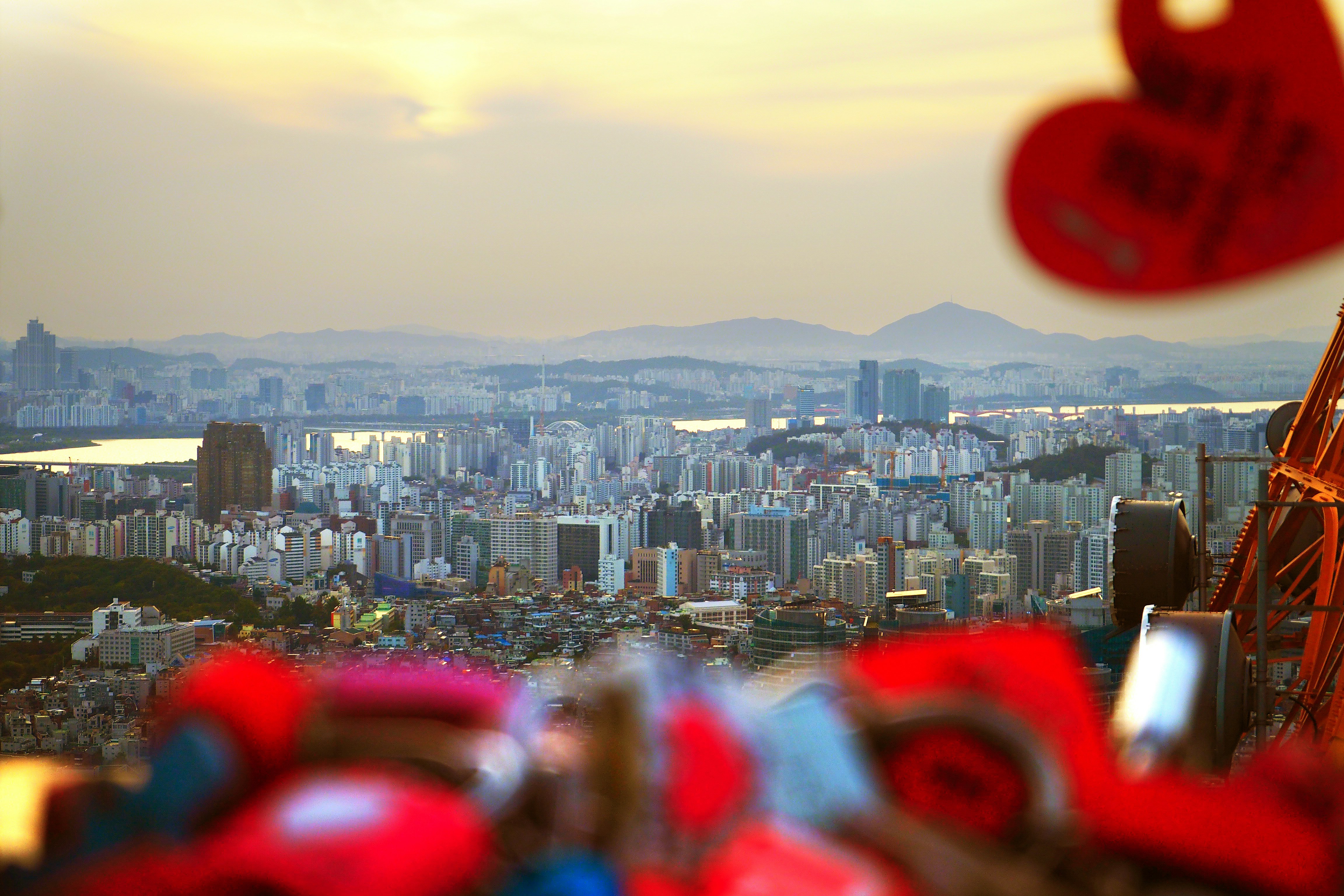 Golden-hour cityscape photographed from a hillside, with the skyline across a harbor and distant hills; foreground red lanterns are blurred to frame the scene.