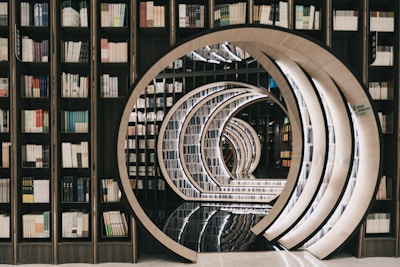 A library with bookshelves filled with various books forms a tunnel-like perspective with circular, mirrored arches. The arches create an illusion of infinity, with the polished floor reflecting the intricate architectural design.