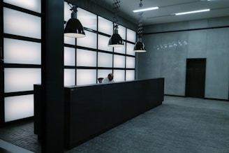 A modern and minimalistic reception area featuring a dark, sleek front desk. A person is seated behind the desk, working under four large, black pendant lights. The backdrop consists of a grid-like pattern of illuminated panels, creating a futuristic atmosphere. The floor is composed of gray tiles, complementing the overall aesthetic.