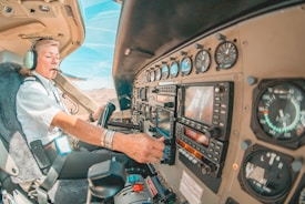 A pilot is seated in the cockpit of an aircraft, wearing a headset and uniform with visible epaulettes. The cockpit features various instruments, gauges, and controls on the dashboard. The pilot appears focused on operating the aircraft, surrounded by a warm interior with a view of the clear sky and landscape through the aircraft window.