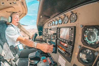 A pilot is seated in the cockpit of an aircraft, wearing a headset and uniform with visible epaulettes. The cockpit features various instruments, gauges, and controls on the dashboard. The pilot appears focused on operating the aircraft, surrounded by a warm interior with a view of the clear sky and landscape through the aircraft window.