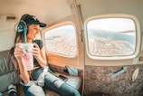 A person is sitting inside a small aircraft, wearing a headset, and holding a frothy pink drink. The individual looks outside the window, enjoying the view of a sprawling cityscape below. Sunlight fills the cabin, creating a warm and inviting atmosphere.