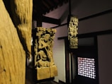 Wooden sculptures featuring intricate carvings of figures and elaborate designs are positioned within a dimly lit interior. The sculptures are mounted on wooden pillars against a backdrop of traditional architecture, with a decorative lattice window on one side.