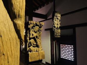 Wooden sculptures featuring intricate carvings of figures and elaborate designs are positioned within a dimly lit interior. The sculptures are mounted on wooden pillars against a backdrop of traditional architecture, with a decorative lattice window on one side.