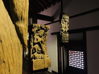 Wooden sculptures featuring intricate carvings of figures and elaborate designs are positioned within a dimly lit interior. The sculptures are mounted on wooden pillars against a backdrop of traditional architecture, with a decorative lattice window on one side.