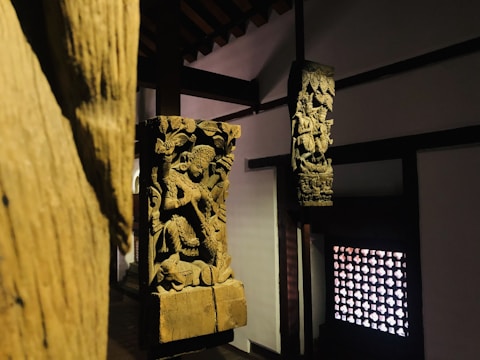 Wooden sculptures featuring intricate carvings of figures and elaborate designs are positioned within a dimly lit interior. The sculptures are mounted on wooden pillars against a backdrop of traditional architecture, with a decorative lattice window on one side.