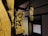 Wooden sculptures featuring intricate carvings of figures and elaborate designs are positioned within a dimly lit interior. The sculptures are mounted on wooden pillars against a backdrop of traditional architecture, with a decorative lattice window on one side.