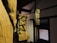 Wooden sculptures featuring intricate carvings of figures and elaborate designs are positioned within a dimly lit interior. The sculptures are mounted on wooden pillars against a backdrop of traditional architecture, with a decorative lattice window on one side.