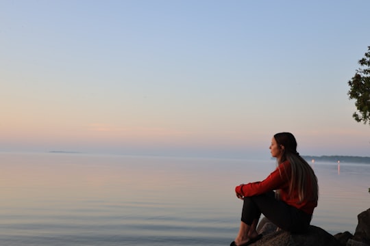 man in red hoodie sitting on rock near body of water during daytime in Barrie Canada