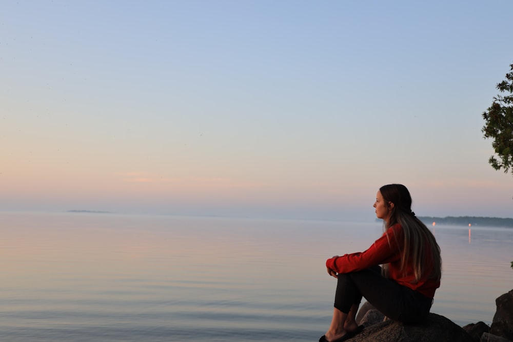 man in red hoodie sitting on rock near body of water during daytime