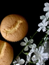 Two golden-brown muffins are placed on a dark background, accompanied by delicate white flowers. The muffins display a crumbly texture, and the flowers have soft petals.