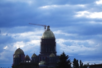 A large domed building under construction, topped with scaffolding and a crane. The sky is filled with thick, dramatic clouds in various shades of blue. Trees and smaller domed structures are visible in the foreground.
