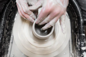 Hands shaping a clay pot on a spinning wheel, dusted with soft clay powder.