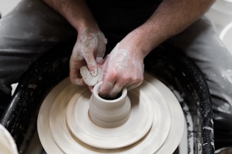 Hands covered in clay are skillfully shaping a pot on a pottery wheel. The person is focused on molding the clay into a symmetrical form, with the wheel in motion and clay scraps scattered around.