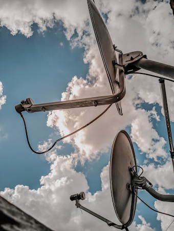 Two satellite dishes are positioned against a backdrop of a bright blue sky with fluffy white clouds. The angle is looking upwards, capturing the curvature of the dishes and the intricate mechanical structures holding them in place.