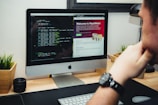 A person is sitting at a desk, looking at an iMac computer. The screen displays a split view with lines of code on the left and a website called 'ReptiMate' on the right, featuring a gecko image. Two small planters with greenery are placed on either side of the monitor, and a camera lens is on the left. The desk has a light wooden surface.