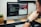 A person is sitting at a desk, looking at an iMac computer. The screen displays a split view with lines of code on the left and a website called 'ReptiMate' on the right, featuring a gecko image. Two small planters with greenery are placed on either side of the monitor, and a camera lens is on the left. The desk has a light wooden surface.