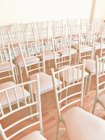 Several empty chairs are arranged in rows on a wooden floor. The chairs have a light cream color and are neatly aligned in a well-lit room.
