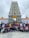 A large group of people is posing in front of a tall and intricately adorned temple tower. The temple architecture features detailed carvings and a central doorway decorated with a yellow and orange drape. The group consists of individuals in casual attire, suggesting a communal or group visit.