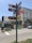 A street corner with a tall signpost displaying multiple colorful signs pointing in different directions, featuring various business names and logos. The background shows a building with an awning, a palm tree, and some pedestrians on the sidewalk. A white truck is parked on the street.