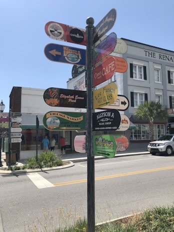 A street corner with a tall signpost displaying multiple colorful signs pointing in different directions, featuring various business names and logos. The background shows a building with an awning, a palm tree, and some pedestrians on the sidewalk. A white truck is parked on the street.
