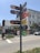 A street corner with a tall signpost displaying multiple colorful signs pointing in different directions, featuring various business names and logos. The background shows a building with an awning, a palm tree, and some pedestrians on the sidewalk. A white truck is parked on the street.