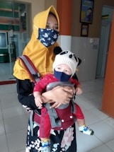 A person wearing a yellow headscarf and a floral face mask is holding a baby. The baby is dressed in a red outfit and is secured in a baby carrier. The baby is also wearing a panda-themed beanie and a face mask. The setting appears to be indoors, likely in a building with tiled floors and some visible decor and signage on the walls.