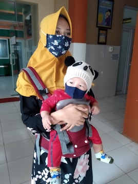 A person wearing a yellow headscarf and a floral face mask is holding a baby. The baby is dressed in a red outfit and is secured in a baby carrier. The baby is also wearing a panda-themed beanie and a face mask. The setting appears to be indoors, likely in a building with tiled floors and some visible decor and signage on the walls.