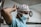 A healthcare worker, wearing a light blue surgical gown, a white mask, and a white cap with red polka dots, adjusts her headwear in a hospital setting. The background features a wall clock and a white and gray interior.