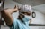 A healthcare worker, wearing a light blue surgical gown, a white mask, and a white cap with red polka dots, adjusts her headwear in a hospital setting. The background features a wall clock and a white and gray interior.