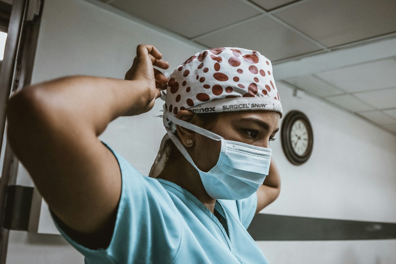 A health worker in a surgical cap tying on a face mask