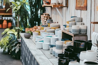 A variety of ceramic pots and vases are arranged on wooden shelves in an outdoor shop setting. The pottery features different patterns and colors. Lush green plants are visible in the surroundings, enriching the natural and artisanal ambiance.