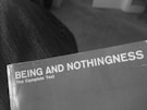 A close-up of the corner of an open book with a plain cover titled 'BEING AND NOTHINGNESS The Complete Text'. The surface appears slightly worn, indicating some use. It rests on a textured fabric surface, likely denim.