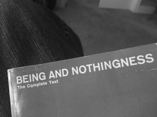 A close-up of the corner of an open book with a plain cover titled 'BEING AND NOTHINGNESS The Complete Text'. The surface appears slightly worn, indicating some use. It rests on a textured fabric surface, likely denim.