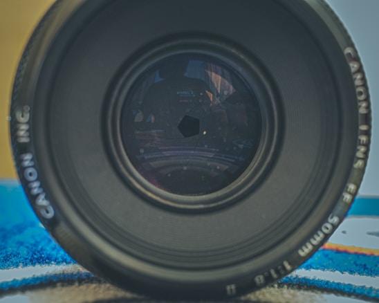 A close-up view of a camera lens displaying intricate details like the aperture blades and inner reflections. The lens branding is visible on the outer ring and it is placed on a surface that has a mix of colors in the background.