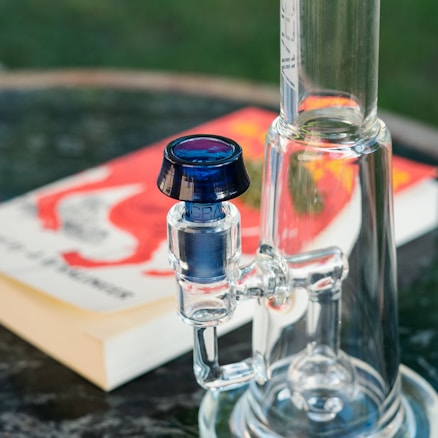A close-up of a glass bong with a blue accented mouthpiece, placed on a table. In the background, a blurred book with a vibrant red and white cover is visible.