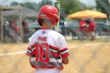 A young baseball player stands on a field, holding a bat and wearing a helmet. The player is seen from the back, with a jersey labeled 'Page' and the number '10'. The setting appears to be a sunny day with a baseball game in progress, and there is a fence in the foreground and other spectators or players in the background.