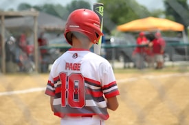 A young baseball player stands on a field, holding a bat and wearing a helmet. The player is seen from the back, with a jersey labeled 'Page' and the number '10'. The setting appears to be a sunny day with a baseball game in progress, and there is a fence in the foreground and other spectators or players in the background.