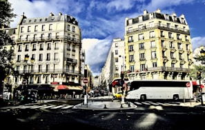 A bustling urban street scene featuring classic Parisian-style buildings with ornate facades and balconies on either side. A large white bus is prominently parked on the right side of the road. Several people are visible walking along the sidewalk, and red awnings can be seen over storefronts. The sky is partly cloudy, and the shadows indicate afternoon lighting.