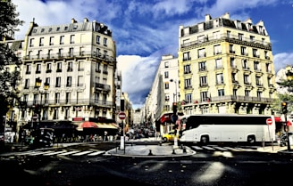 A bustling urban street scene featuring classic Parisian-style buildings with ornate facades and balconies on either side. A large white bus is prominently parked on the right side of the road. Several people are visible walking along the sidewalk, and red awnings can be seen over storefronts. The sky is partly cloudy, and the shadows indicate afternoon lighting.