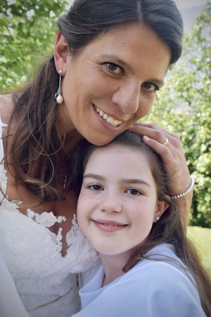 A woman and a young girl are captured in a close embrace outside in a leafy setting. The woman smiles warmly, resting her head gently on the girl, who has a content expression. The woman is wearing a lace-trimmed garment and pearl earrings, with her dark hair falling naturally over her shoulders. The girl has long hair and is dressed in a simple white outfit.