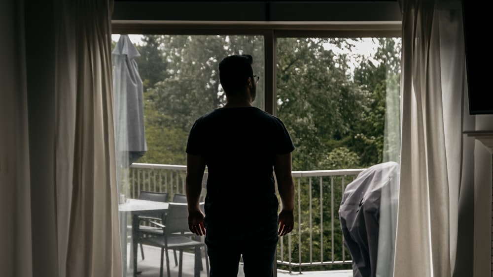 Hombre en camiseta negra de cuello redondo de pie cerca de la cortina blanca de la ventana