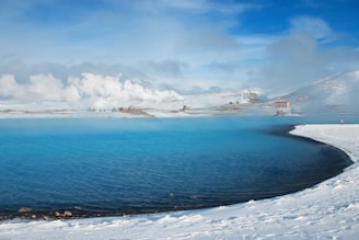 A serene landscape featuring a vibrant blue geothermal lagoon surrounded by snow-covered land. Billowing clouds of steam rise from the water, with distant hills and sparse buildings visible under a bright blue sky.