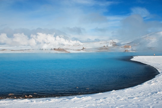 A serene landscape featuring a vibrant blue geothermal lagoon surrounded by snow-covered land. Billowing clouds of steam rise from the water, with distant hills and sparse buildings visible under a bright blue sky.