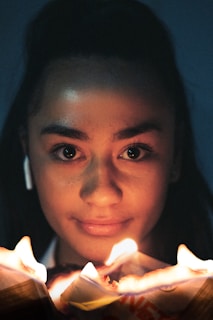 A person holding a burning piece of paper close to their face, creating a dramatic glow that highlights their facial features. The background is dark, enhancing the brightness of the flames.